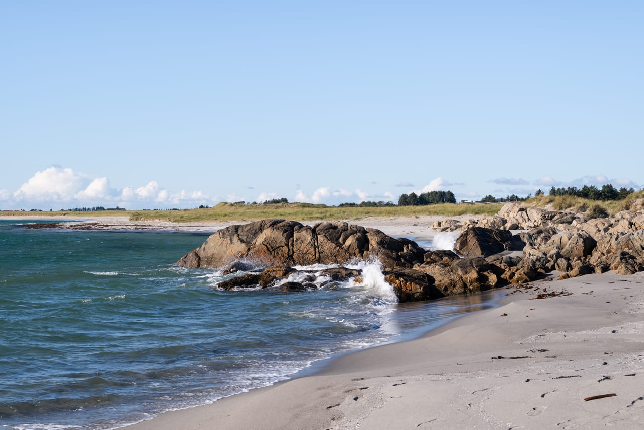 Haviksanden på Lista, med sandstrand og klipper der bølgene bryter mot steinene, under blå himmel med noen hvite skyer.