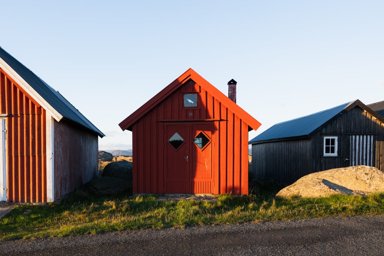 Lite, rødt naust med rombeformede vinduer i døra, mellom to andre naust i Verevågen, i lavt kveldslys.