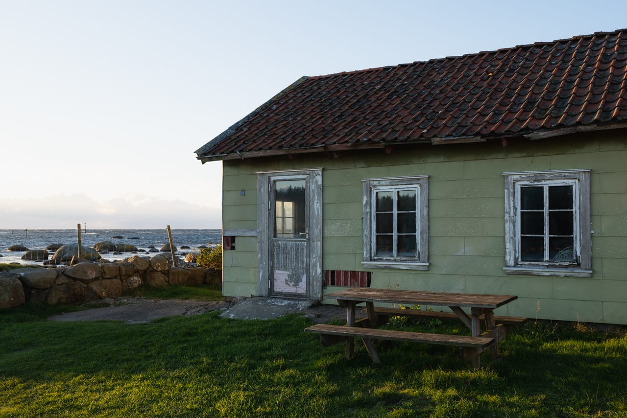 Lite, grønt hus i Verevågen med teglsteinstak, gamle vinduer og et slitt benkebord på plenen, med en steinmur og havet bak.