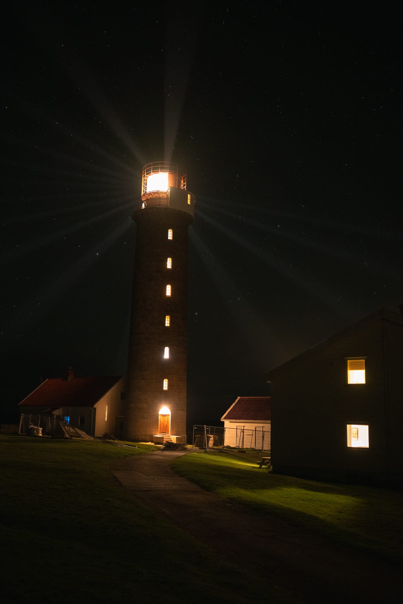 Lista fyr om natta, med lysstråler fra lykta, stjernehimmel og opplyste vinduer i bygningene rundt.