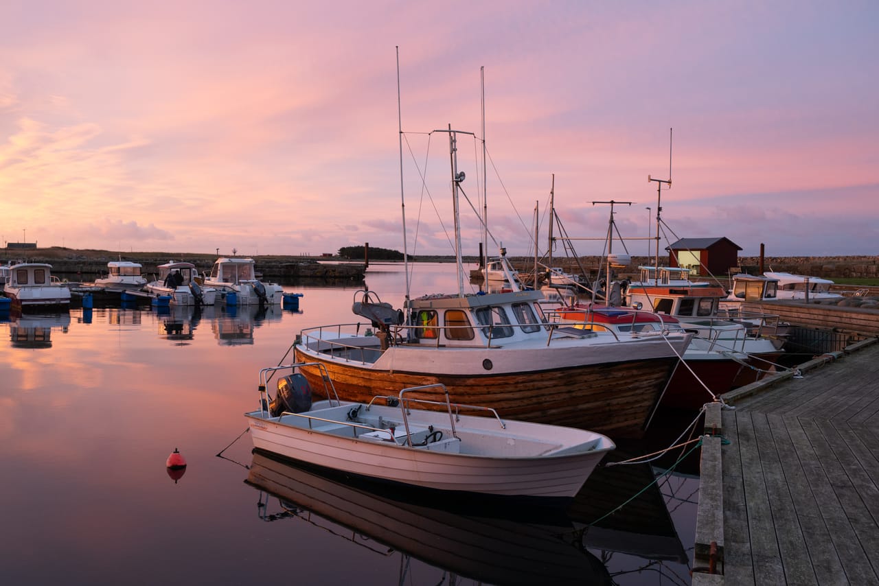 Fiskebåter fortøyd ved en trebrygge i Borshavn i soloppgangen, med rosa og fiolett himmel som speiler seg i det stille vannet.