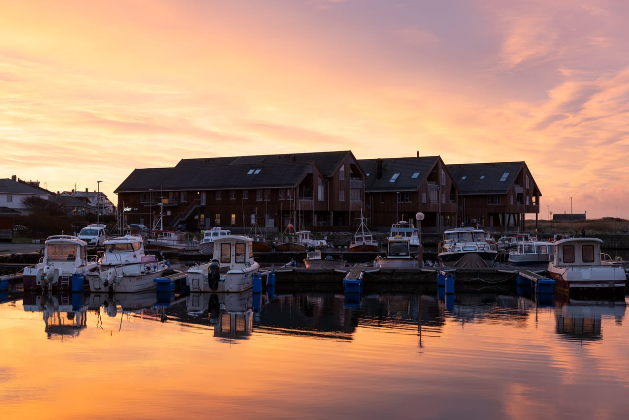 Borshavn i soloppgangen, med småbåter ved flytebrygga og brune bygninger langs havna under en glødende oransje og rosa himmel, speilet i vannet.