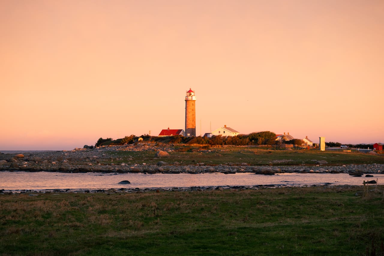 Lista fyr med hvite og røde hus rundt, i varmt morgenlys over en stille vik med rullestein, sett fra et jorde.