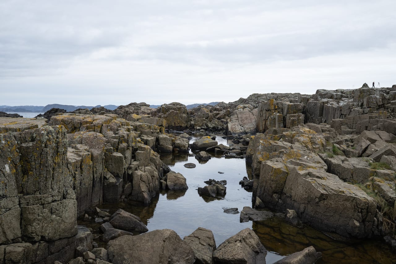 En smal kløft med stille vann mellom kantete bergvegger ved Håle, med en fotograf og et stativ ved en varde oppe på kanten, og fjell i det fjerne.