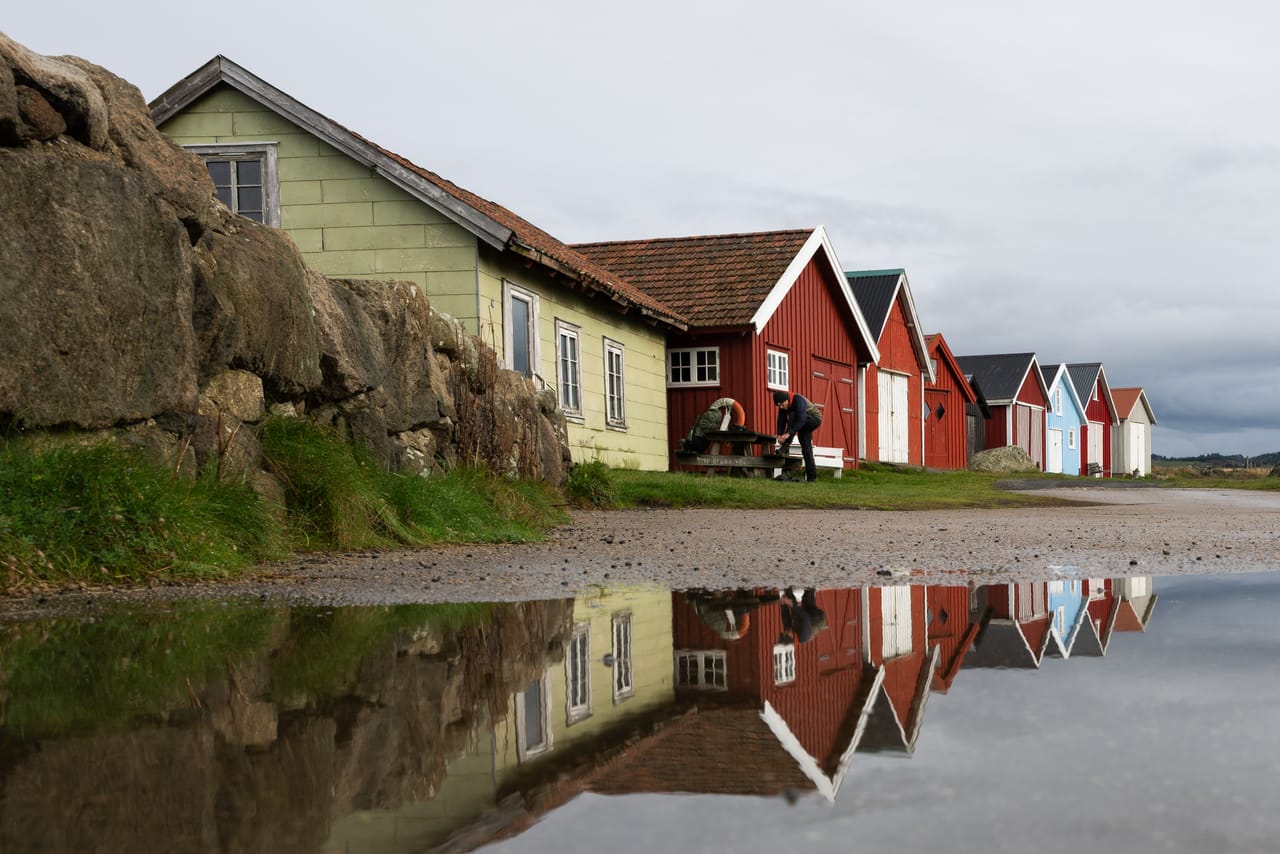 Naustrekken i Verevågen speilet i en stor vanndam på grusveien, med et grønt hus, røde og blå naust og en person ved et benkebord.