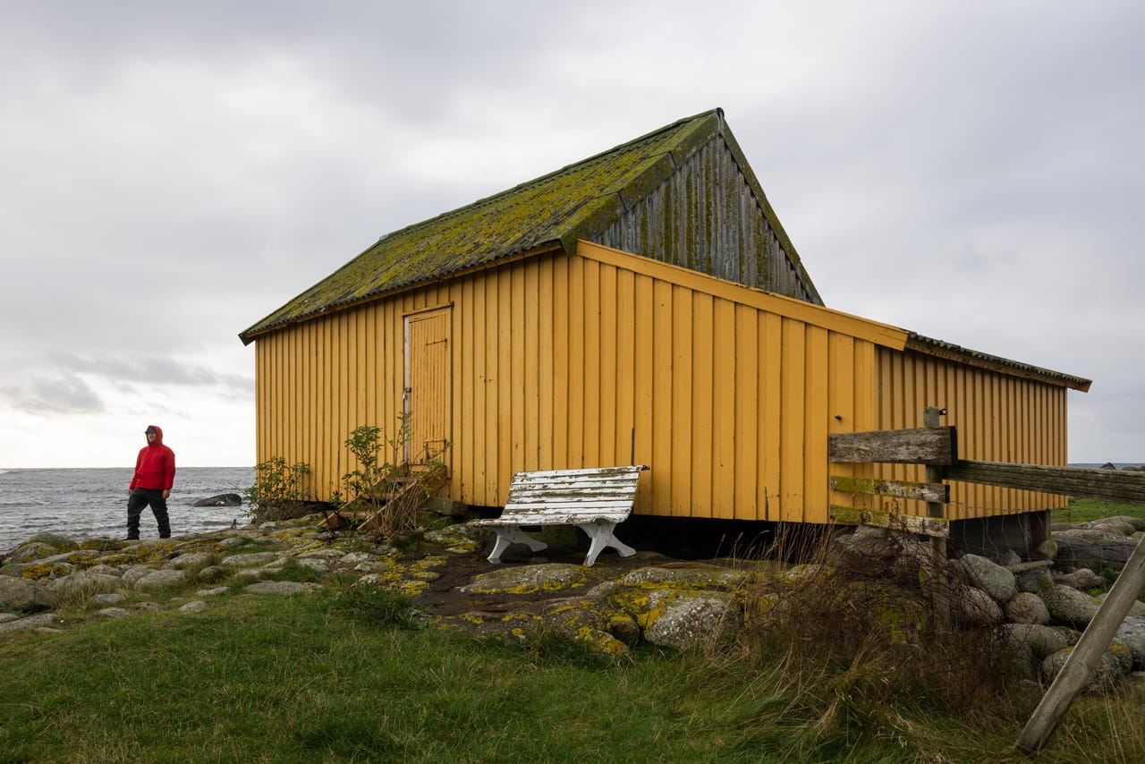 Gulebua, det gule naustet ved Lista fyr, med mosegrodd tak og en hvit benk foran, under grå skyer, med en person i rød jakke nede ved sjøen.