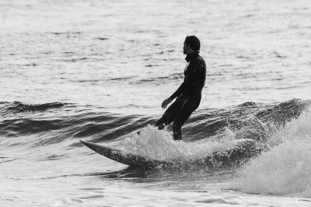 Surfer i våtdrakt nesten i silhuett, stående på brettet i en liten bølge mot glitrende vann, i svart-hvitt.