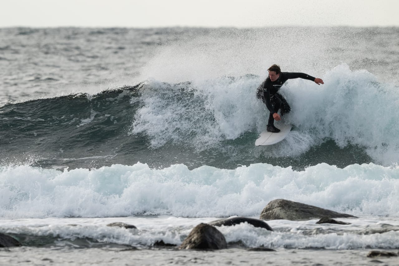 Surfer i våtdrakt som svinger på et hvitt surfebrett i en bølge som brekker ved Lista fyr, med skum og steiner i forgrunnen.