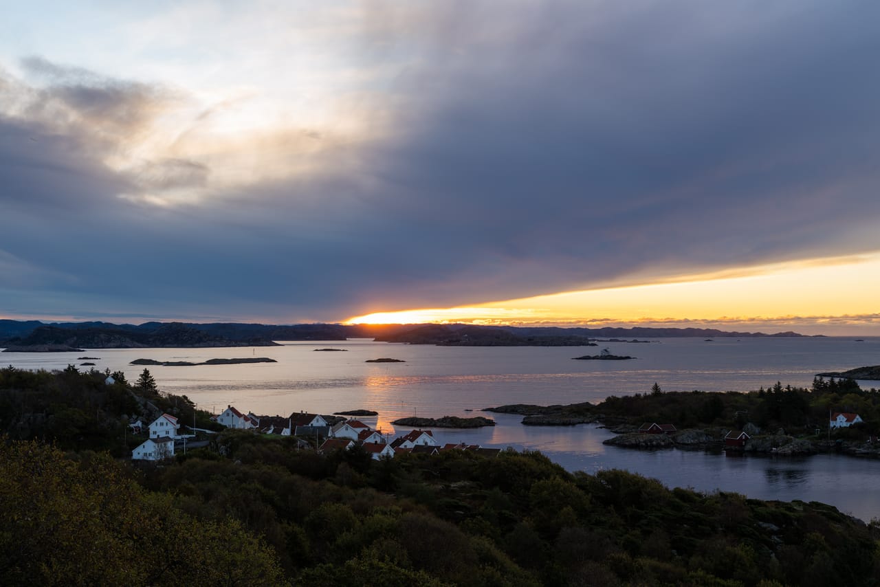 Soloppgang over skjærgården ved Loshavn, med en smal, gyllen stripe under et blågrått skydekke og hvite hus med røde tak ved sjøen nedenfor.