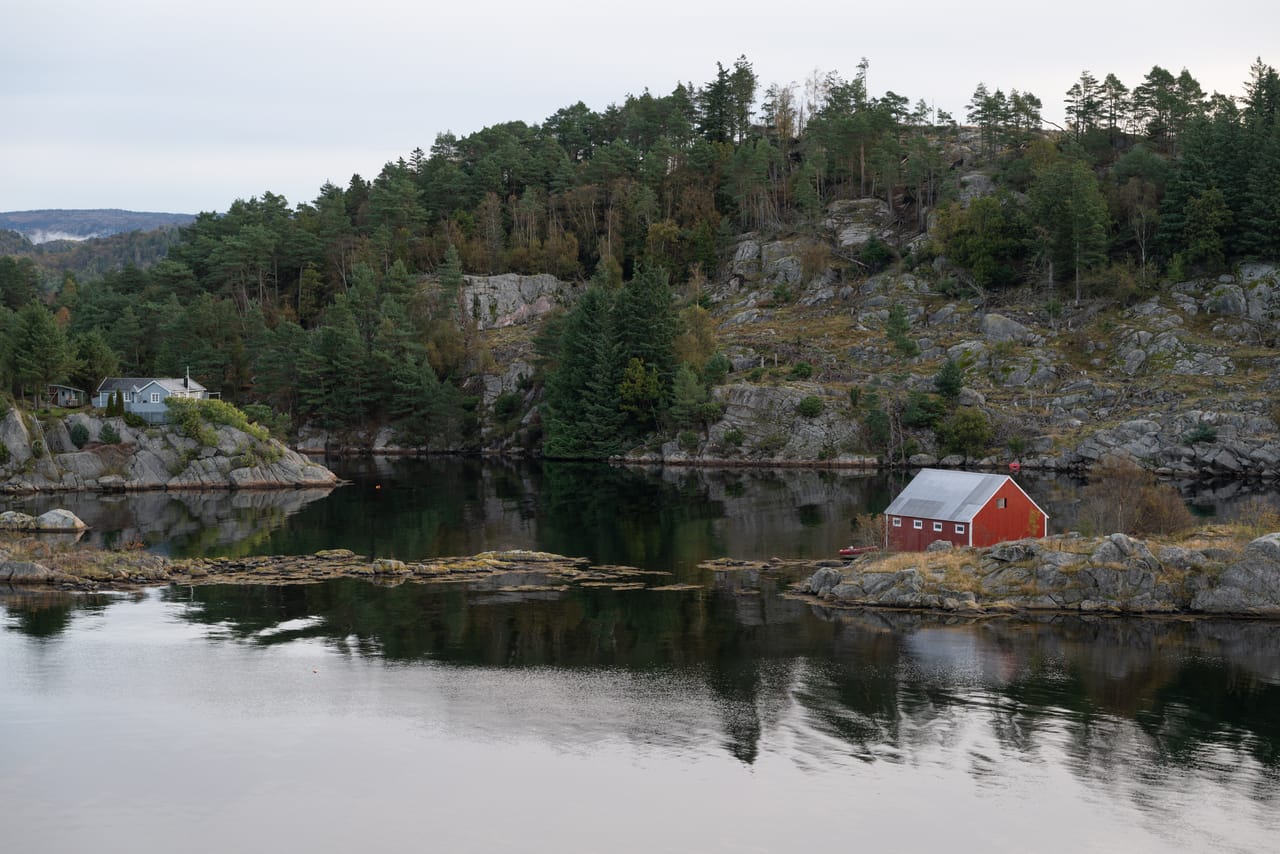 Stille bukt ved Loshavn som speiler furuskog og grå bergknauser, med et rødt naust på en lav knaus og et hus i skogkanten, under en grå himmel.