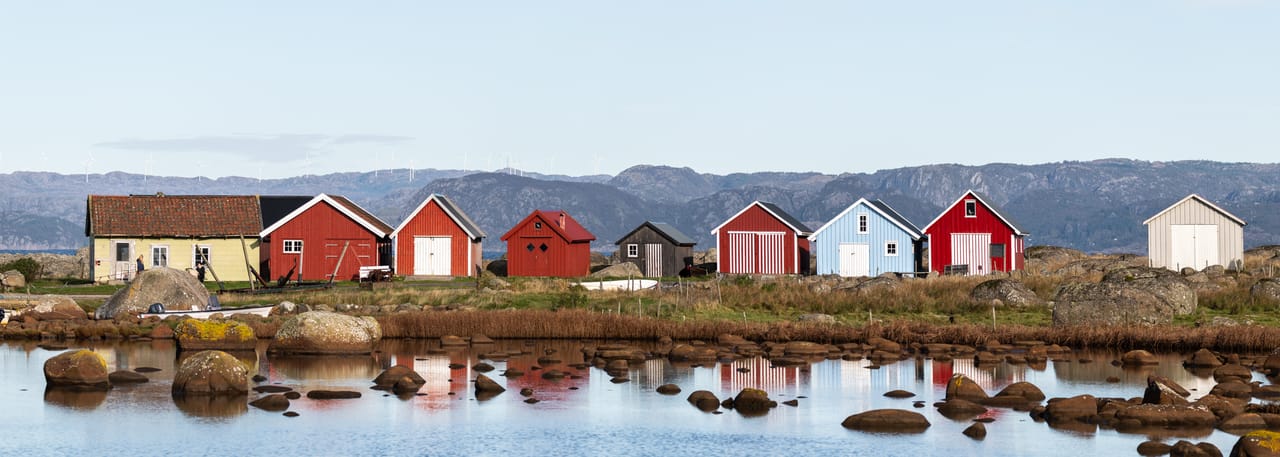 Naustrekken i Verevågen i panorama, med et gult hus og røde, svarte, blå og hvite naust speilet i stille vann mellom steiner, og vindturbiner på fjellene bak.