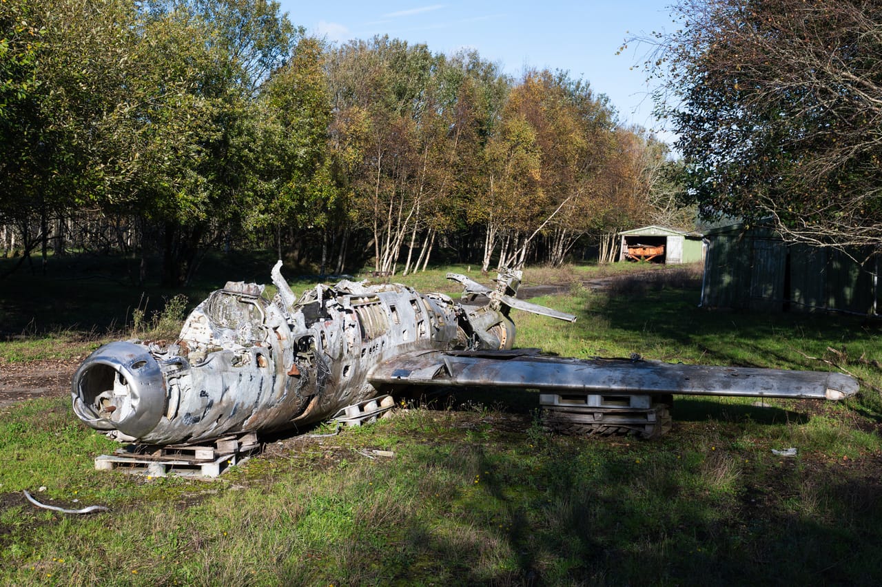Vrak av et eldre jetfly ved flystripa på Lista, med blank, bulkete metallkropp og rundt luftinntak i nesen, på paller i gresset foran høstfargede trær.