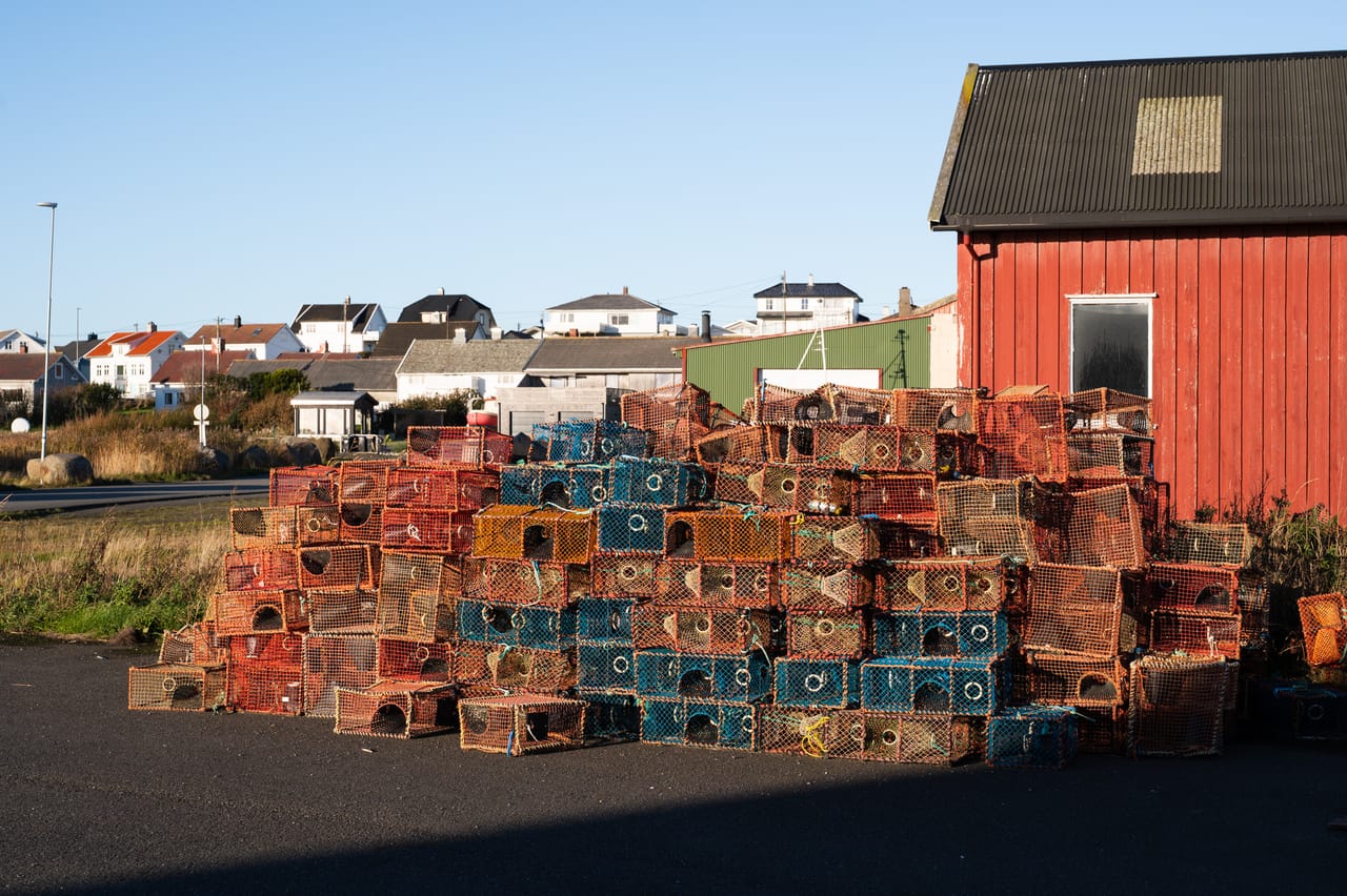 Høye stabler med teiner i oransje og blått foran en rød bygning, med hvite hus i Borhaug i bakgrunnen under blå himmel.