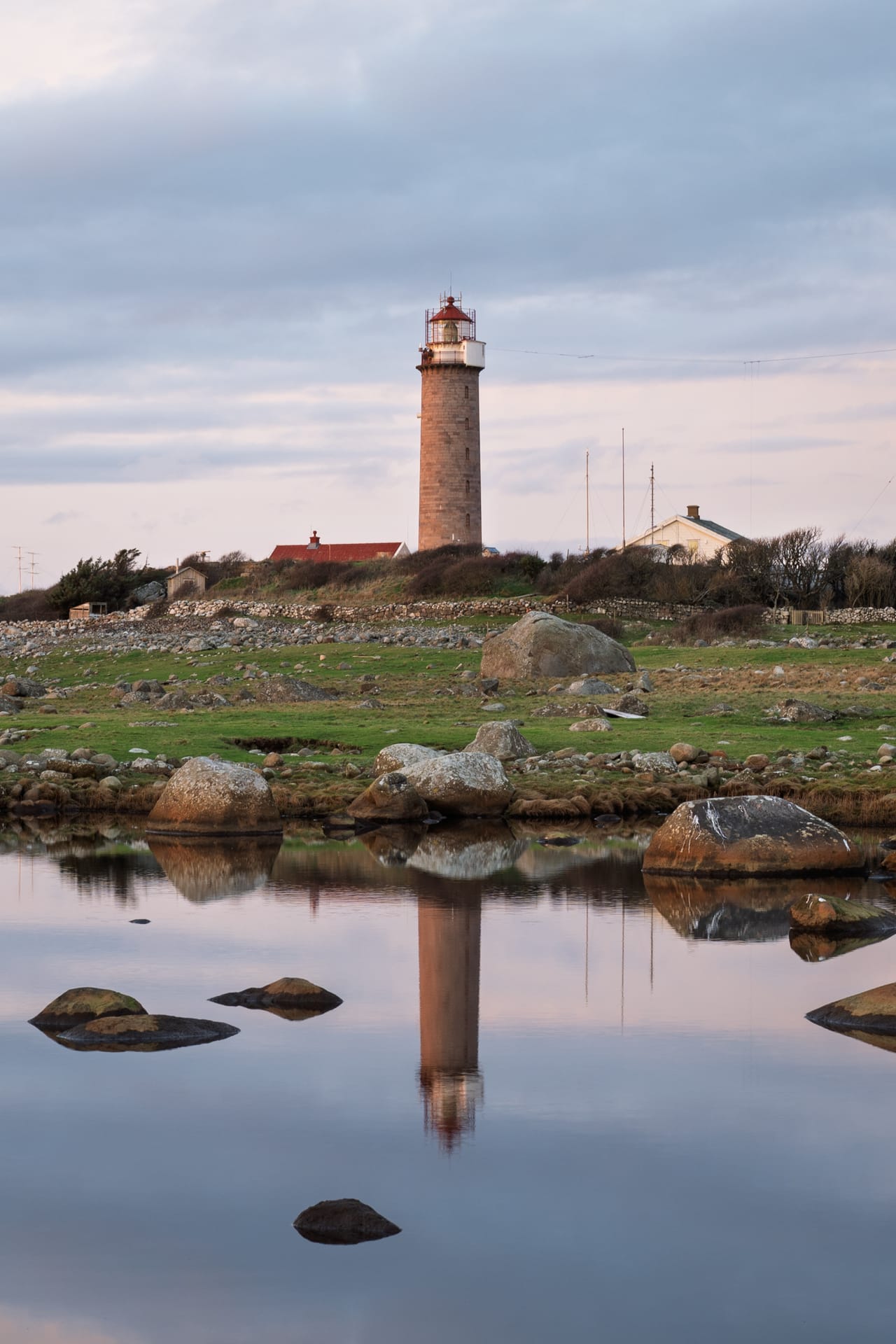 Lista fyr speilet i et rolig vannhull i våtmarka, med stein i forgrunnen og rosa skumringshimmel.