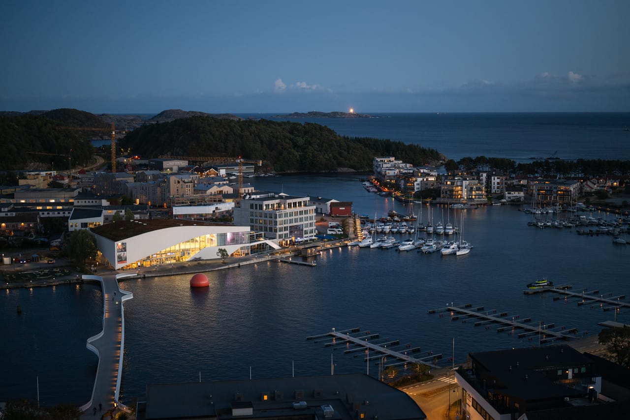 Utsikt over Mandal og havnebassenget ved Buen kulturhus i blåtimen, med et fyrlys synlig langt ute ved horisonten.