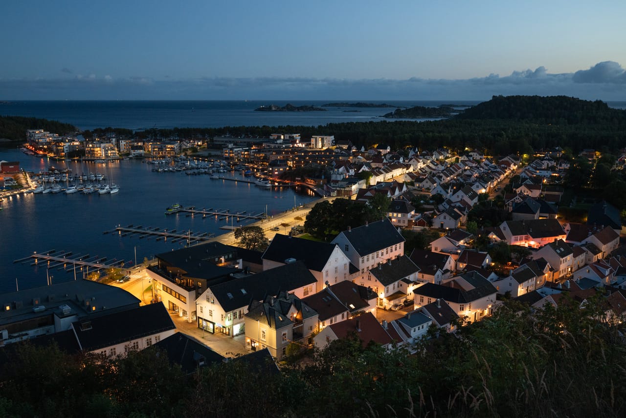 Mandal sentrum sett ovenfra i blåtimen, med gatelys og opplyste hus langs vannkanten.