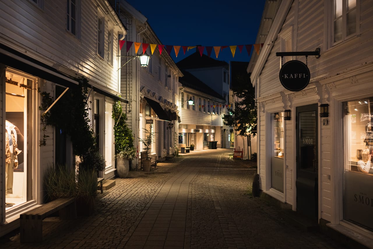 En brosteinsgate mellom hvitmalte hus i Mandal sentrum om kvelden, med vimpler og butikkskilt tent.