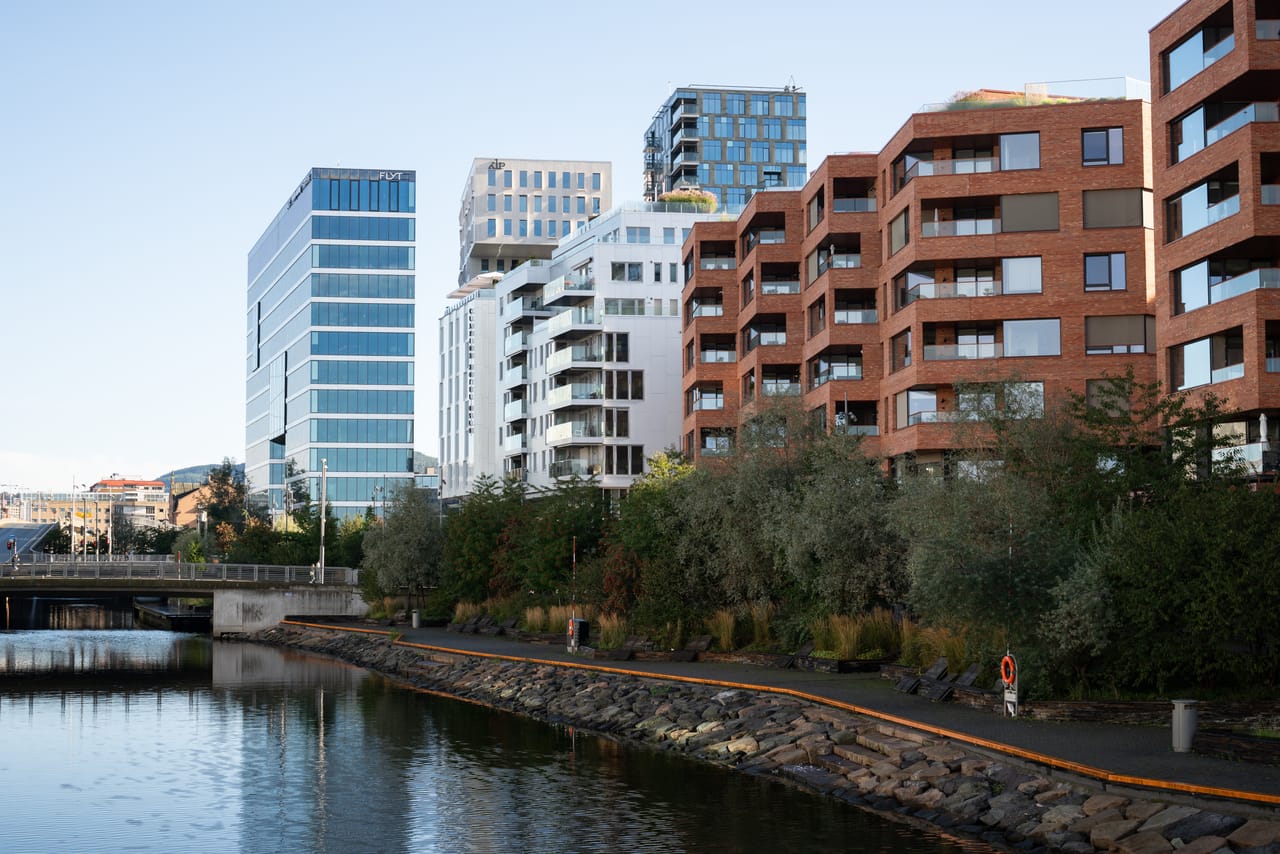 Nybygde boligblokker i tegl og glass langs en kanal, med en gangbro og en steinsatt kaipromenade i forgrunnen.