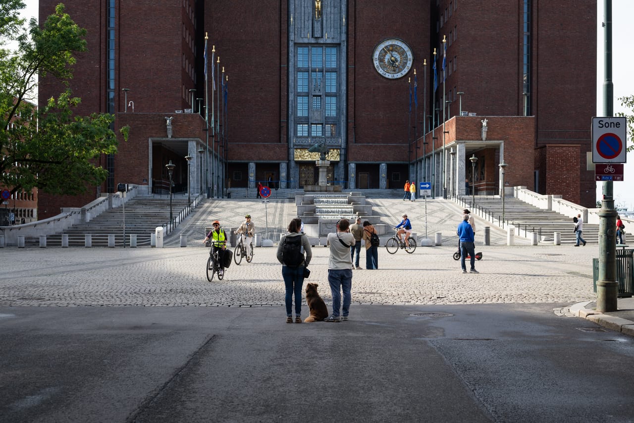 Oslo rådhus sett fra plassen foran, med syklister, fontenen og to personer med hund i forgrunnen.