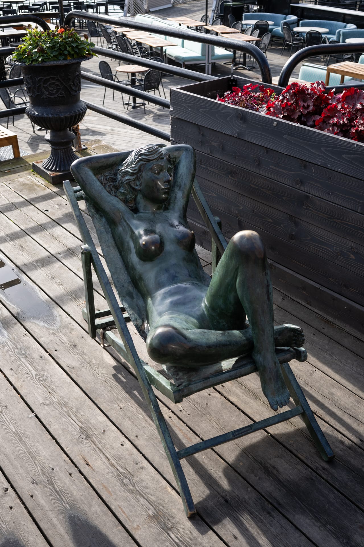 En bronsestatue av en solende kvinne i solstol, på en terrasse med blomsterkasser.