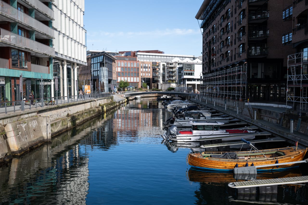 En smal kanal mellom moderne bygninger, med båter fortøyd og en gangbro i bakgrunnen.