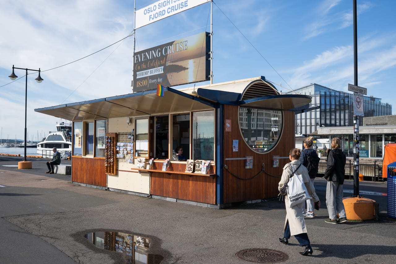 Den gamle billettkiosken for fjordcruise ved Rådhusbrygga, pyntet med regnbueflagg, med Nasjonalmuseet skimtet i bakgrunnen.