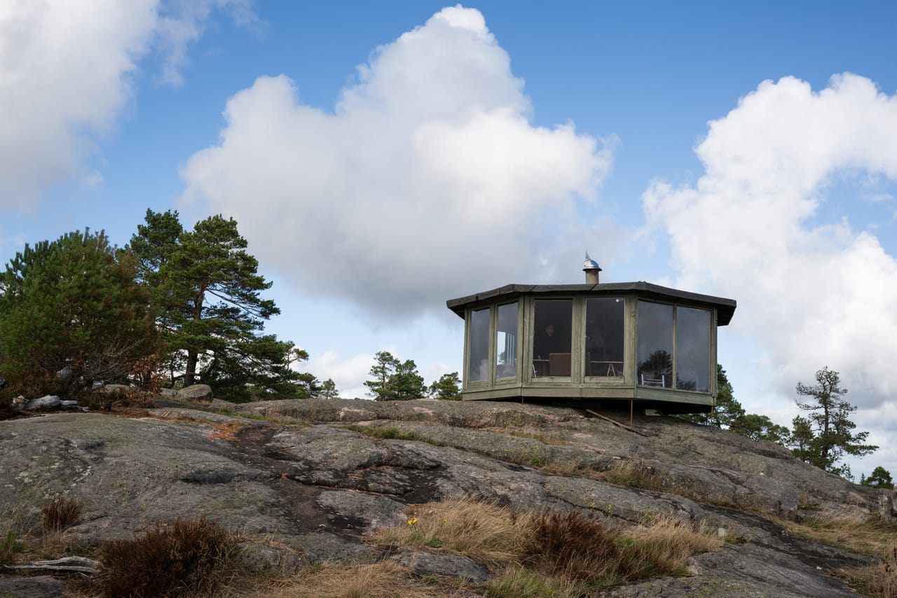 Den åttekantede grillhytta med store vinduer på svaberget på Slettehei, under blå himmel og hvite skyer.