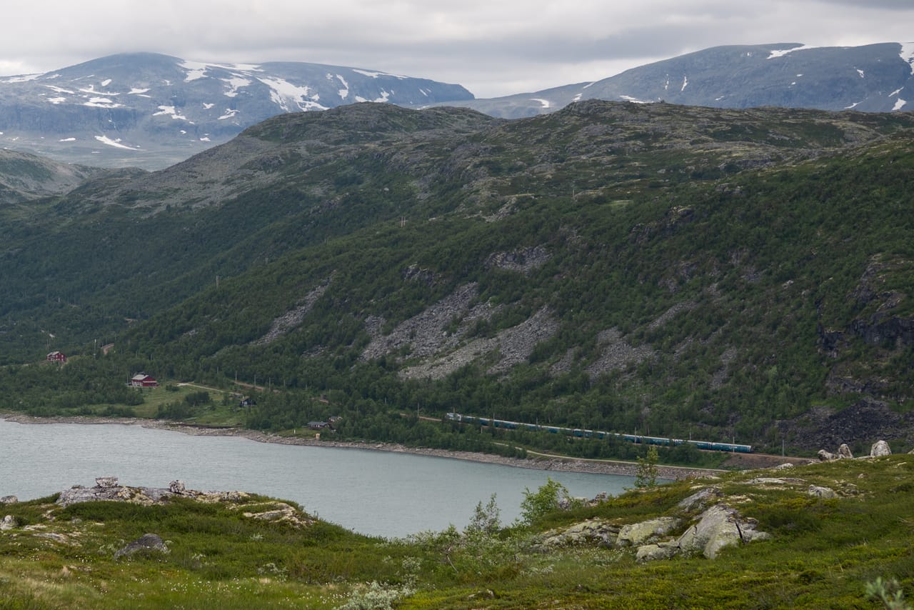 Bergensbanen langs et fjellvann nedenfor en bjørkekledd li, med snøflekkete fjell i bakgrunnen.