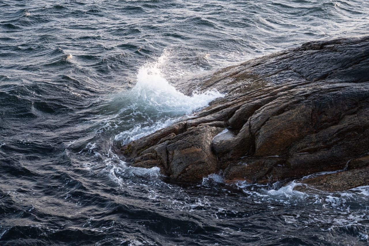 En bølge slår opp i hvitt skum mot et rødbrunt svaberg, med mørkt, urolig hav rundt.