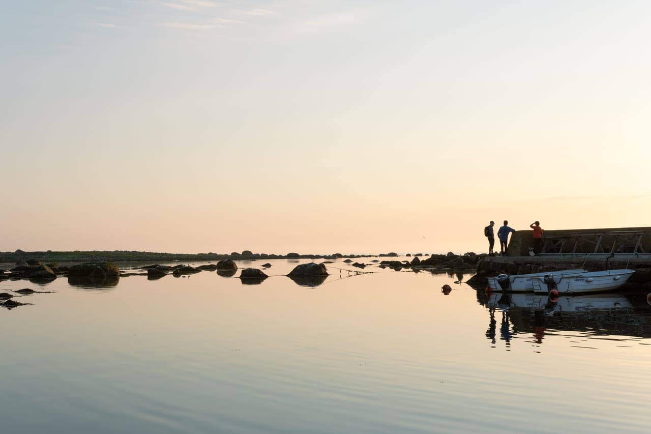 Tre personer i silhuett på moloen i Verevågen på Lista, med en liten båt fortøyd i det speilblanke vannet under en pastellfarget himmel.