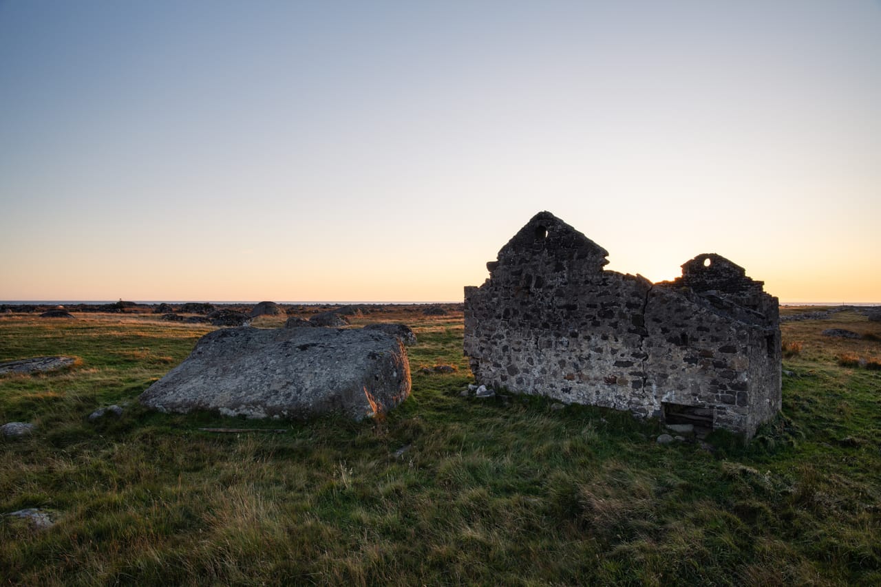 Ruinen av et gammelt steinhus og en stor kampestein i gresslandskapet mellom Lista fyr og Steinodden, i motlys fra solnedgangen.