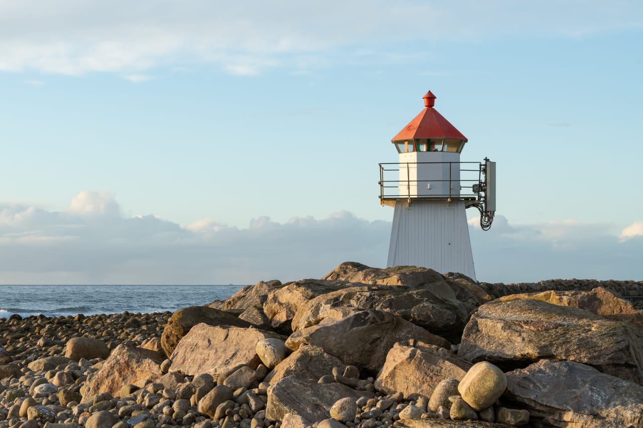 Den hvite fyrlykta med rødt tak på steinmoloen i Borhaug på Lista, med runde rullesteiner i forgrunnen og havet bak i varmt ettermiddagslys.