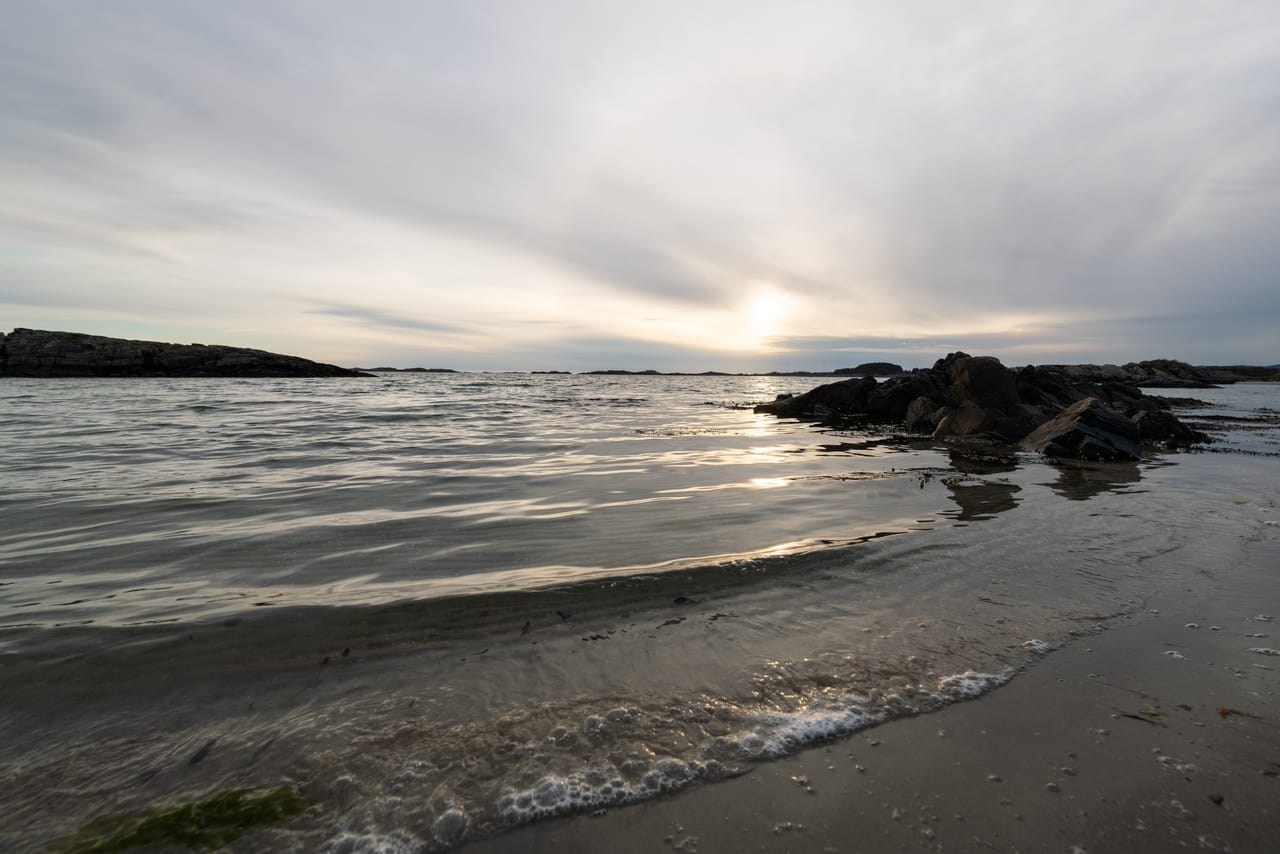 Sola skinner gjennom et tynt skylag over havet ved Einarsneset på Lista, med en liten bølge som renner opp på sandstranden og mørke svaberg bak.