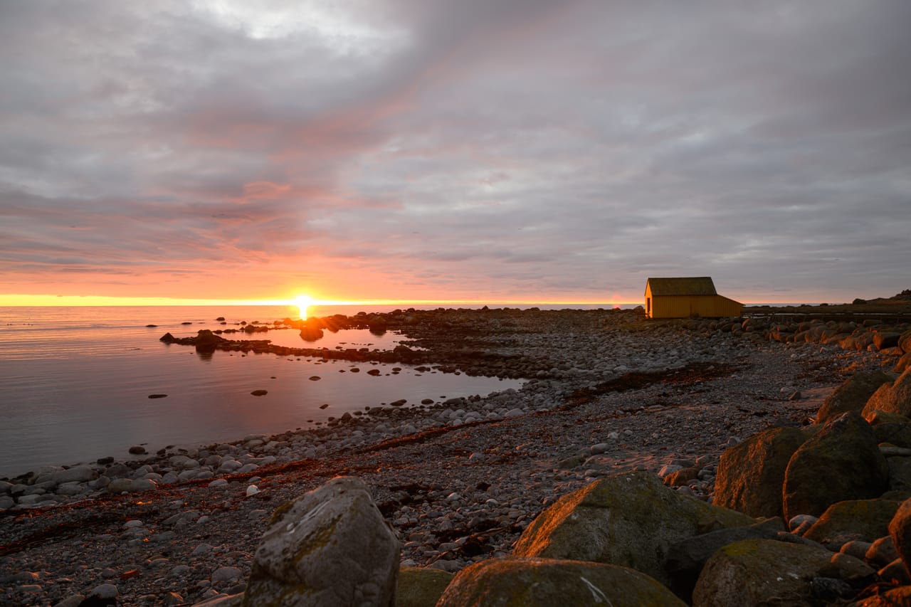 Den gule Gulebua ved strandkanten på Lista mens sola går ned i havet, med steinstrand i forgrunnen under rosa skyer.