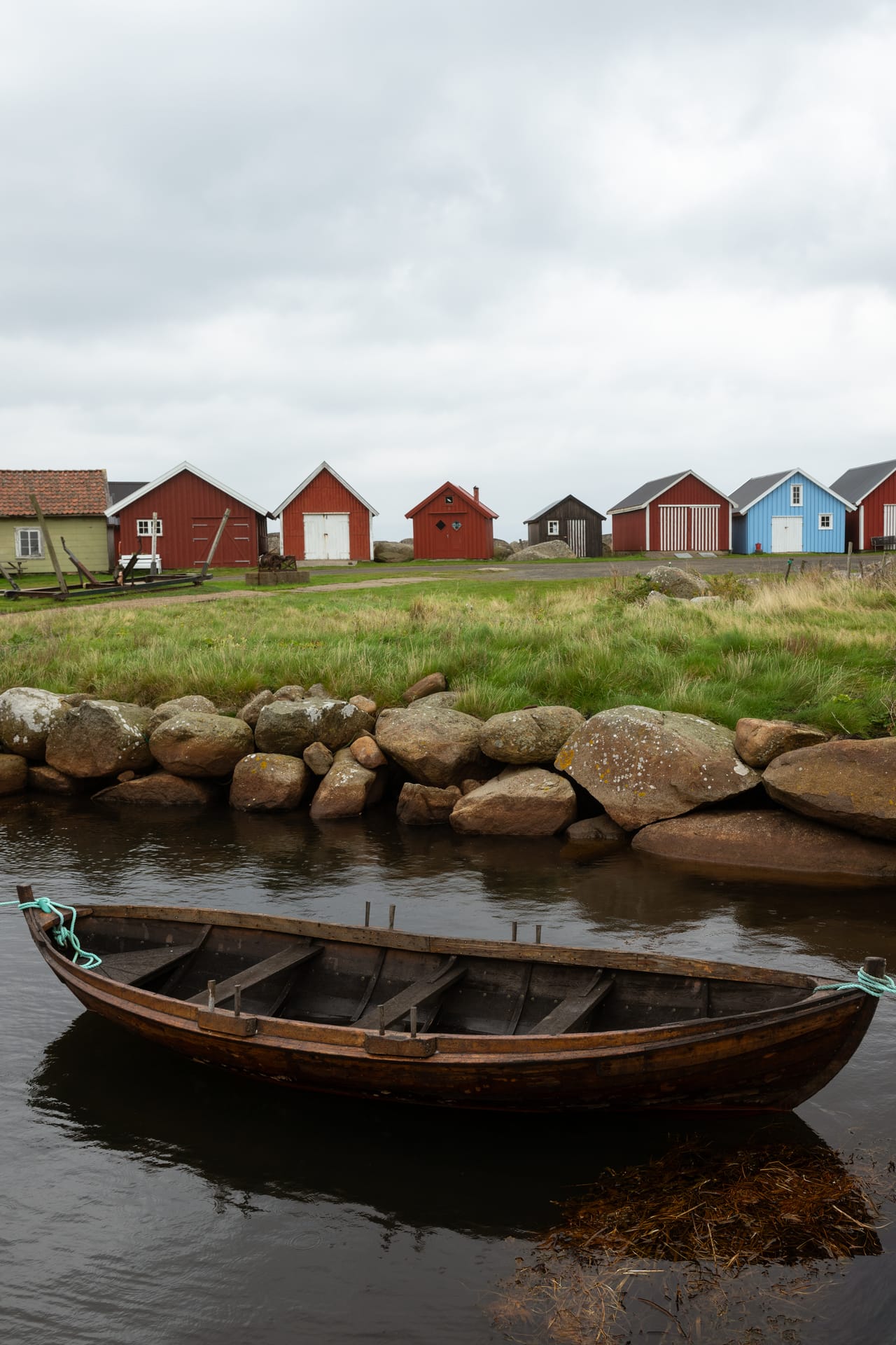 En gammel trebåt ligger fortøyd ved en steinmur i Verevågen på Lista, med en rekke røde, hvite og blå naust bak under gråvær.
