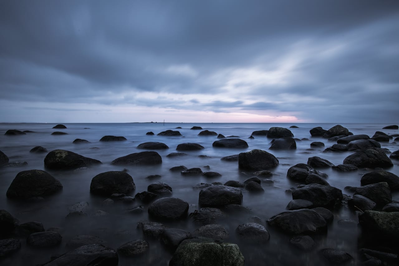 Blanke, mørke rullesteiner i stille vann ved Østhasselstrand på Lista, under et tungt skydekke i skumringen.