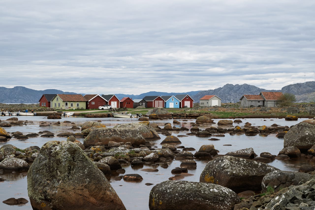 Lavdekte steiner i grunt vann i Verevågen på Lista, med en rekke røde, grønne og blå naust og fjell i bakgrunnen under gråvær.