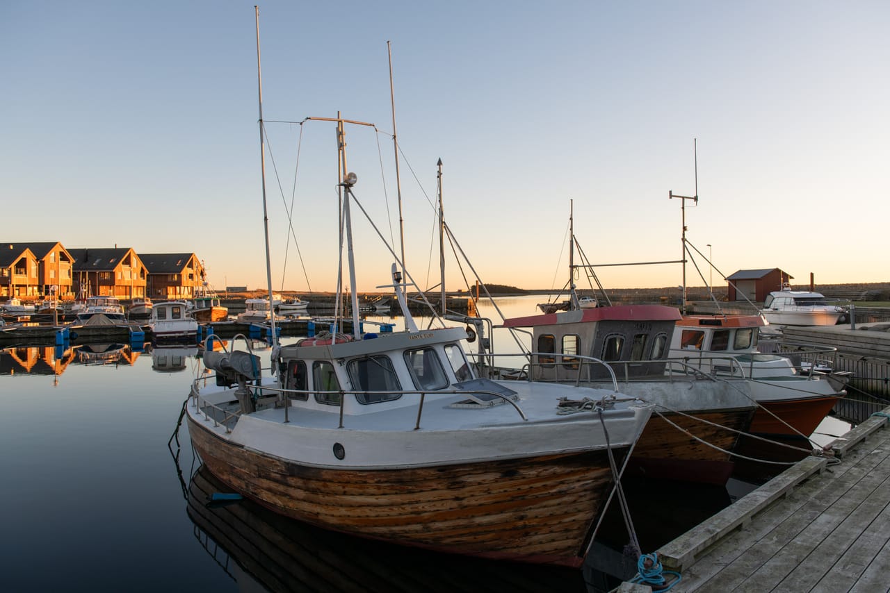 Gamle fiskebåter i tre fortøyd ved trebrygga i Borhaug havn på Lista, med stille vann og lavt sollys på husene bak.