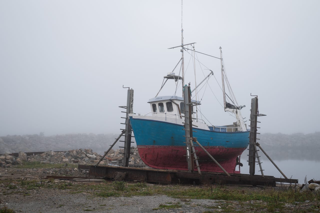 En blå og rød fiskebåt står på slipp på land i Borhaug på Lista, støttet av trestivere i tåka.
