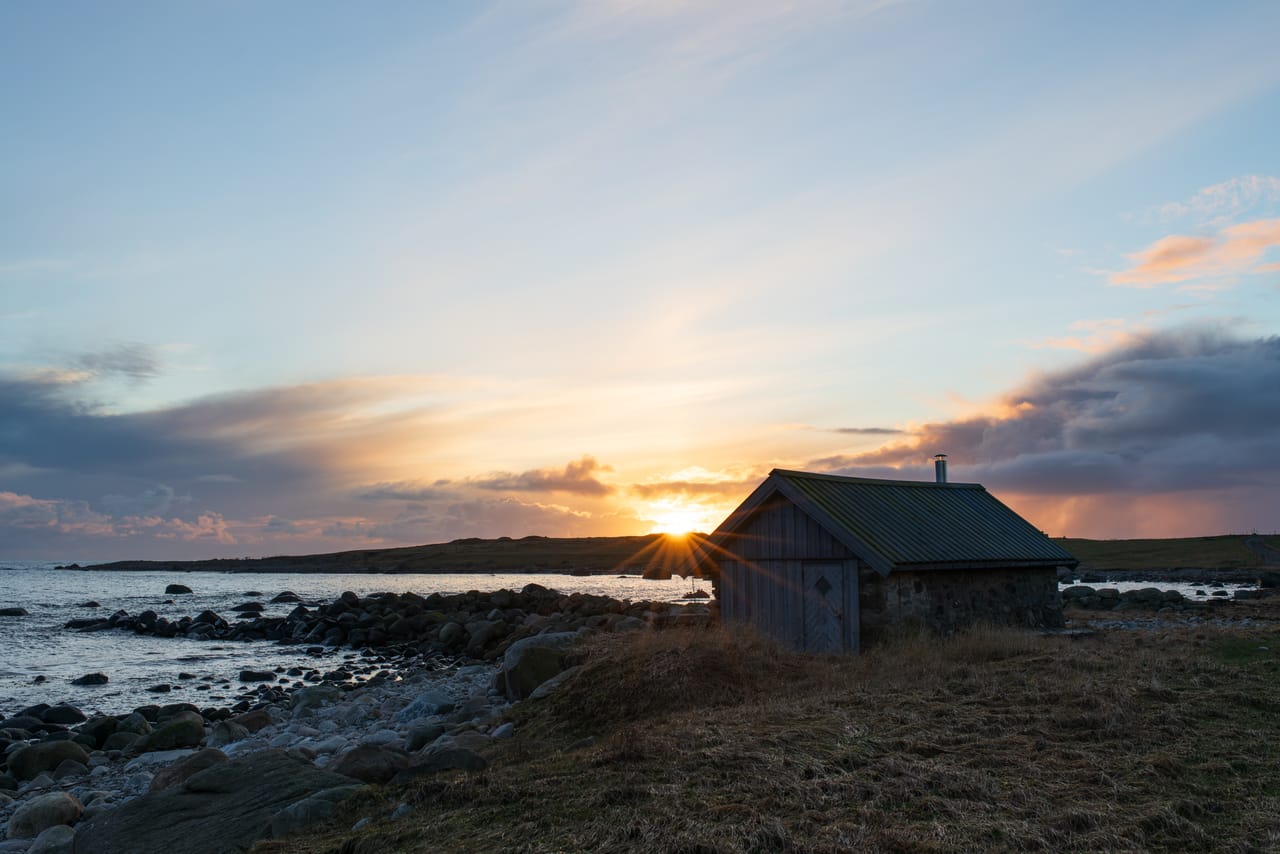 Sola går ned bak en liten bu av stein og tre på Østhasselstrand på Lista, med solstråler, steinstrand og skyer i rosa og blått over havet.