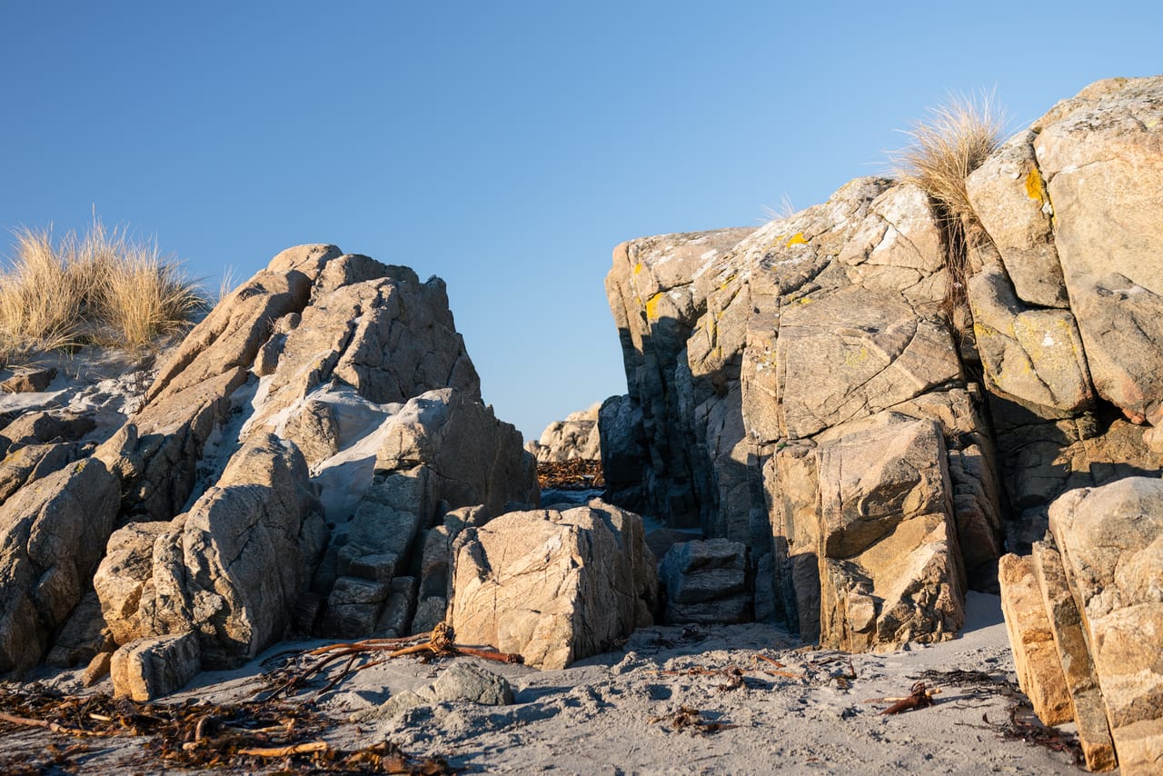 To store steinformasjoner med gul lav og tuer av marehalm rammer inn en smal passasje på Haviksanden på Lista, med tørr tare i sanden og lavt vintersollys.