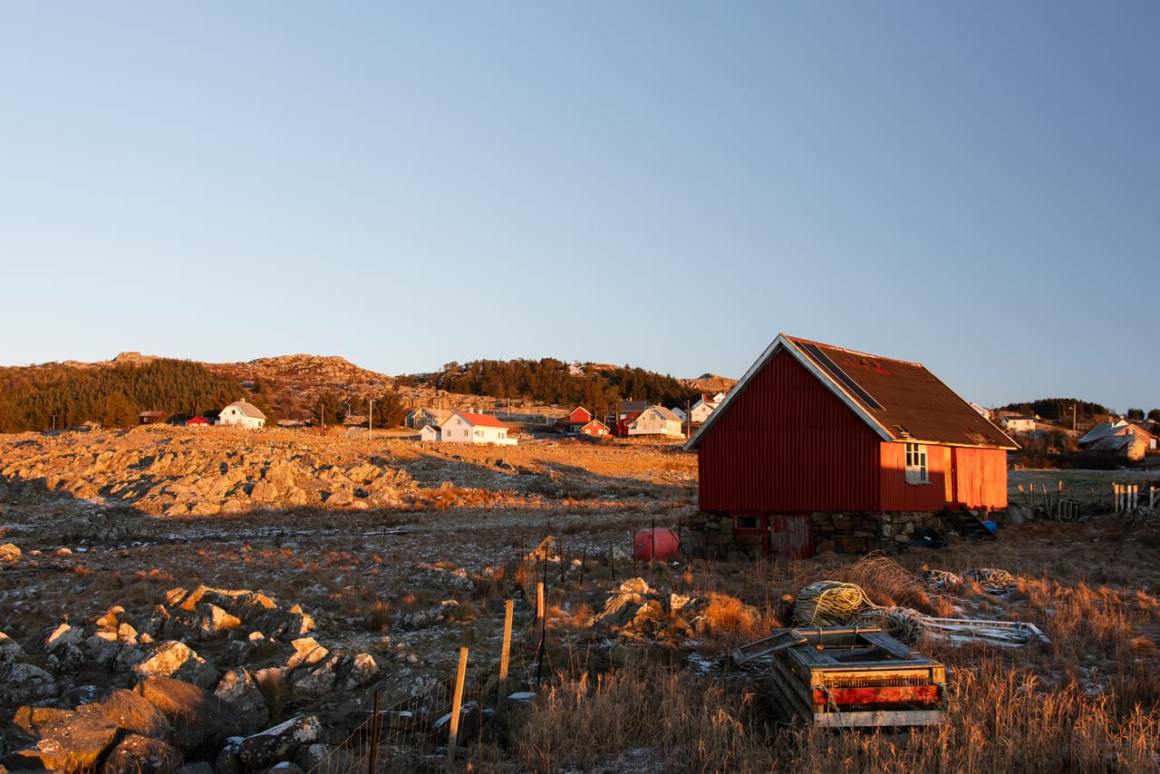 Et rødt trehus på steinmur lyser i lav vintersol på Jøllestø på Lista, med tauverk og fiskeutstyr i det tørre gresset foran og hus spredt i det steinete terrenget bak.