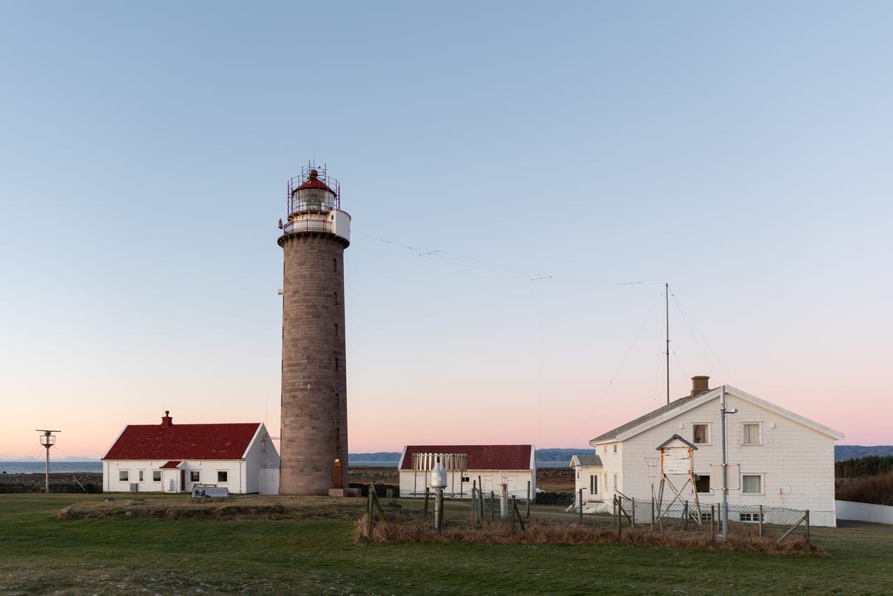 Det murte steintårnet på Lista fyr med rød lyktekuppel, omgitt av hvite fyrbygninger med røde tak, under en rosa og blå kveldshimmel med havet i bakgrunnen.