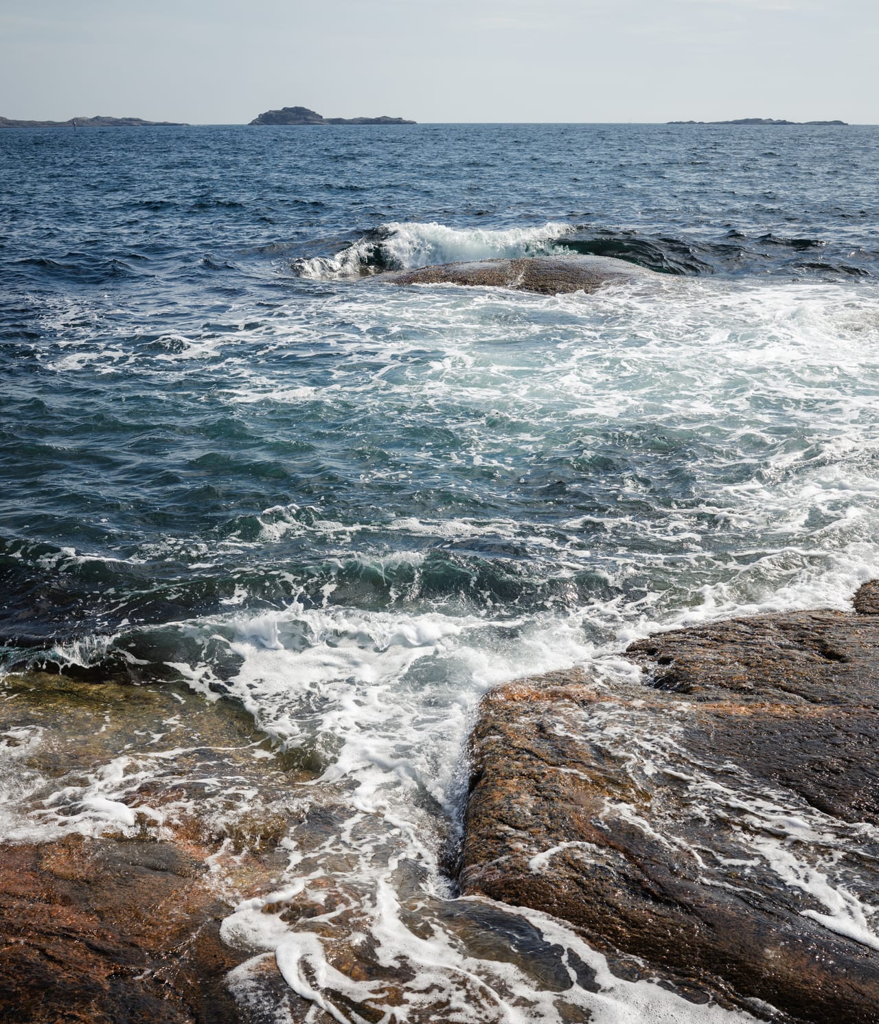 Havet slår innover rødbrune svaberg ved Langenes i Søgne, med skum og lave holmer synlige mot horisonten.
